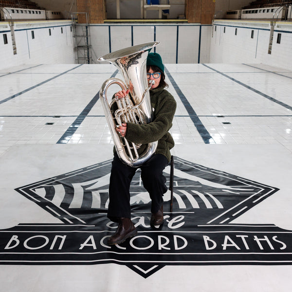 Person holding a large tuba in an indoor pool area with 'Bon Accord Baths' branding.