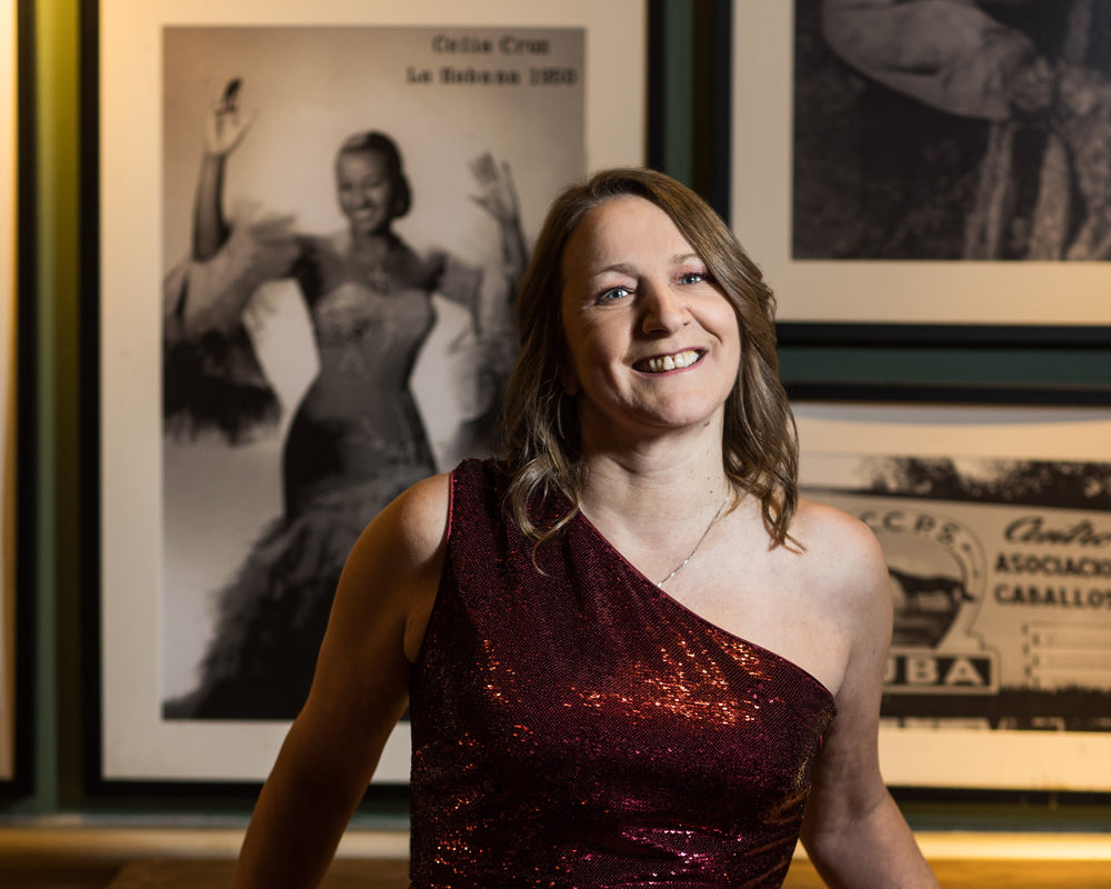 A woman in a red sequined dress standing in front of a wall with framed pictures, smiling at the camera.
